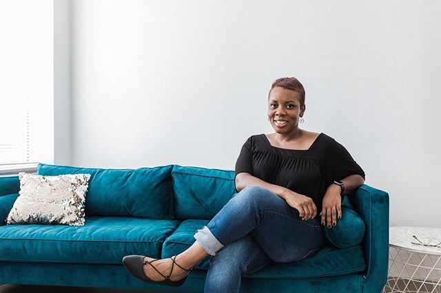 Black girl sitting on turquoise couch. She is smiling looking at the camera, wearing a black shirt, blue jeans, and black flats.