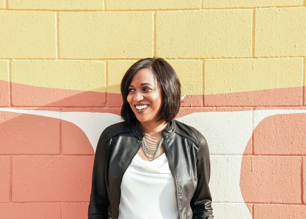Black woman standing in front of a yellow and white mural outside wearing a leather jacket. She is smiling, looking to the left.
