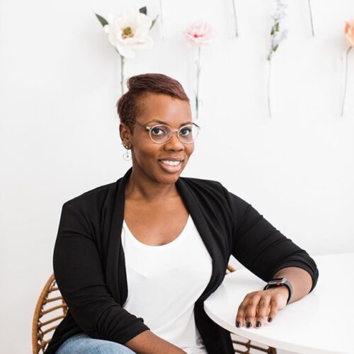 black woman with short hair and glasses sitting at a white table in front of floral background