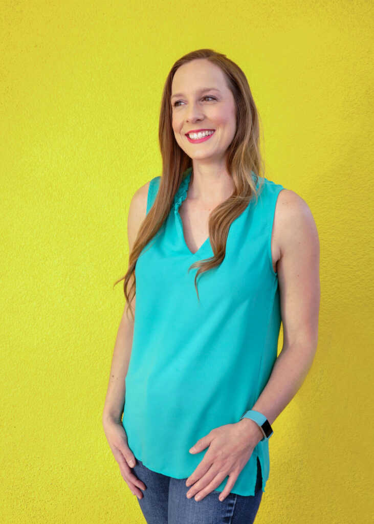 girl with blonde hair standing in front of yellow wall wearing a green sleeveless shirt